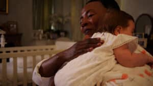 A person gently holds a sleeping baby wrapped in a white blanket over their shoulder, perhaps beginning to help sleep train babies. They are in a softly lit room with a crib and framed photos in the background. Night Nannies London