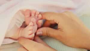 Close-up of a babys tiny feet being gently held by two adult hands, while the baby enjoys peaceful sleep wrapped in a soft white blanket. The adult, with glittery nail polish, embodies tenderness and care in this serene moment. Night Nannies London