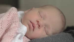 A sleeping newborn baby, 1-3 months old, with eyes closed, wrapped in a soft pink knitted blanket. The babys peaceful expression conveys tranquility and comfort. The softly blurred background highlights the serene face, offering a gentle sense of baby sleep ease. Night Nannies London