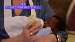 A caregiver in a denim shirt and white apron is bottle-feeding a baby wrapped in a blue blanket. The text Tip 3: Help your 1-3 months old sleep better by ensuring theyre full is shown at the top in a purple box. Night Nannies London
