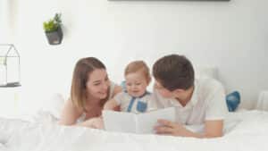 A family enjoys safe sleep as they lie on a bed, reading a book together. A woman and man, both smiling, flank a baby who curiously gazes at the book in the mans hands. The room is bright with white bedding and a plant adorning the wall. Night Nannies London