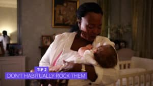 A woman is gently holding a baby in her arms, standing in a room with soft lighting. A purple text box at the bottom says, Tip 2: Encourage Baby to Self-Settle. The setting is warm and homey, with framed pictures on a shelf behind. Night Nannies London