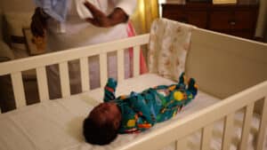 A baby in colorful pajamas lies in a crib, crying softly as they attempt to self-settle. A person beside the crib reaches out a comforting hand towards the baby. A blanket hangs on the cribs side, and a dresser is visible in the background. Night Nannies London