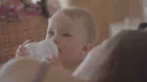 A 6-month-old baby with light hair is drinking from a bottle while reclining against an adult. The background features a decorative basket with pink flowers, creating a soft, warm atmosphere perfect for nurturing and supporting babies in their sleep train routine. Night Nannies London