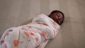 A baby is peacefully sleeping, wrapped in a white blanket adorned with orange flamingo patterns, showcasing the art of swaddling. The tiny bundle rests comfortably on a textured beige surface. Night Nannies London