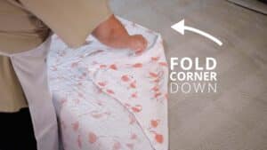 A person is folding down the corner of a floral-patterned bed sheet, much like preparing to swaddle a baby. An arrow points to the folded corner with the text FOLD CORNER DOWN next to it. Night Nannies London