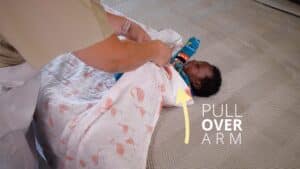 A person demonstrates how to swaddle a baby lying on a soft patterned surface. The baby, in colorful clothing, is nestled snugly. An arrow directs the swaddling technique with the text PULL OVER ARM, guiding this essential parenting step. Night Nannies London