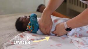 A caregiver demonstrates how to swaddle a baby, wrapping a patterned blanket snugly around the little one and tucking the fabric securely under its body. The infant, dressed in colorful clothing, rests on a changing surface. Text on the image reads Tuck Under, with an arrow highlighting the technique. Night Nannies London