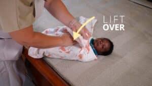 An adult demonstrates how to swaddle a baby by wrapping them in a patterned flamingo swaddle on a bed. A yellow arrow and the words “LIFT OVER” are displayed, guiding the proper completion of this comforting process. Night Nannies London
