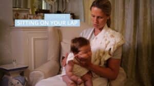 A woman is seated in a chair, holding a baby on her lap. The baby is sitting forward, supported by the womans hands to help wind your baby. Text on the image reads, Position 1: Sitting on your lap. The cozy room adds warmth to this tender moment. Night Nannies London