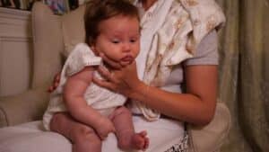 A baby in a white outfit is gently held by an adult seated on a chair, with a careful hand supporting the little ones face. A patterned cloth is draped over their shoulder like a gentle breeze, while the indoor setting features curtains softly framing this tender moment. Night Nannies London