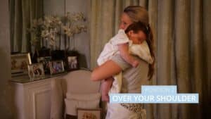 A woman helps wind her baby by gently holding them over her shoulder in a cozy room adorned with family photos and flowers on a cabinet. The text Position 2: Over Your Shoulder is displayed on the image. Night Nannies London